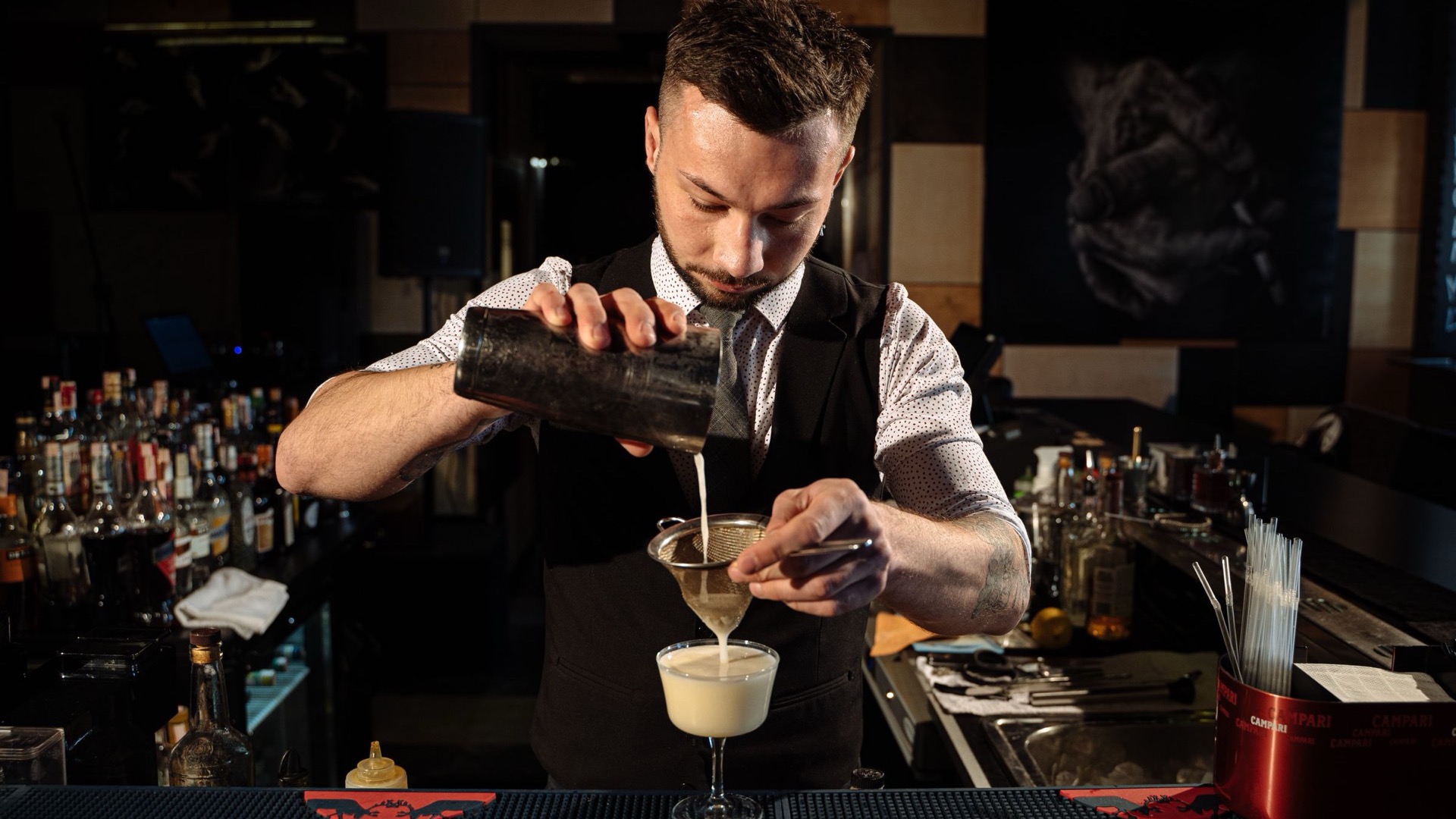A bartender straining a cocktail into a glass at a bar counter.