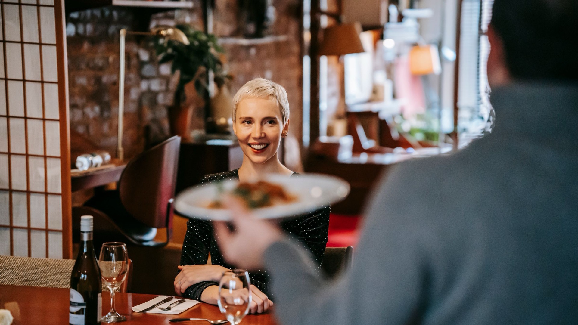 Positive couple having date in bright restaurant while man serving plate with food to woman sitting in chair at table with wine bottle and glasses near cutlery with napkin