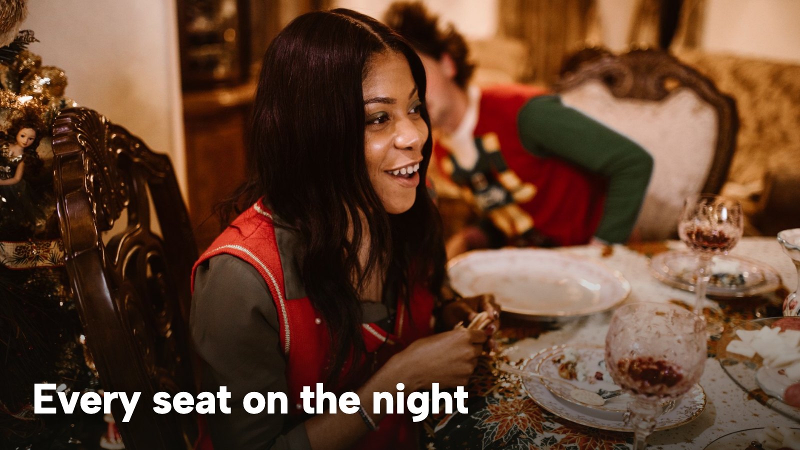 A smiling guest in a festive jumper sits at a Christmas dinner table with plates and glasses. The image carries the words "Every seat on the night".