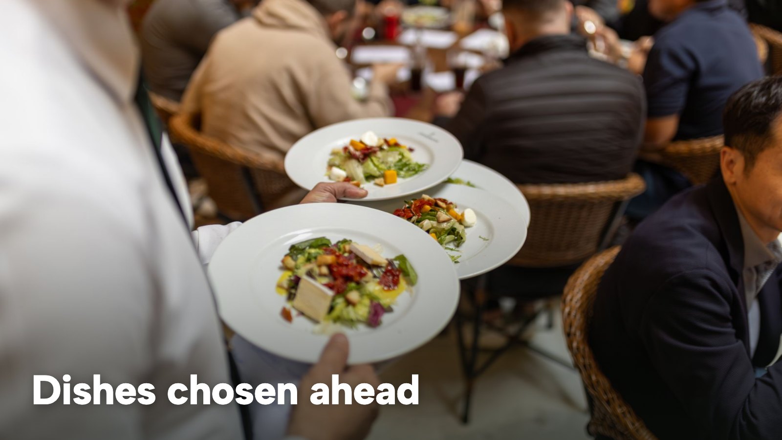 A server carries three plated dishes to a full restaurant table. The image carries the words "Dishes chosen ahead".