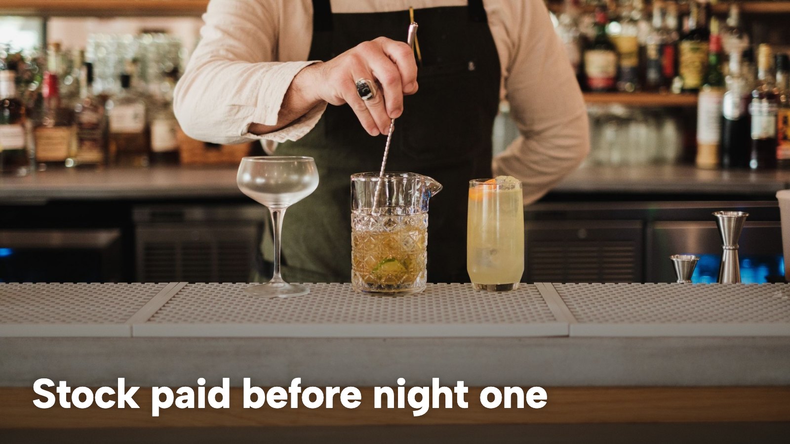 A bartender stirring a cocktail at a well-stocked bar. The image carries the words "Stock paid before night one".