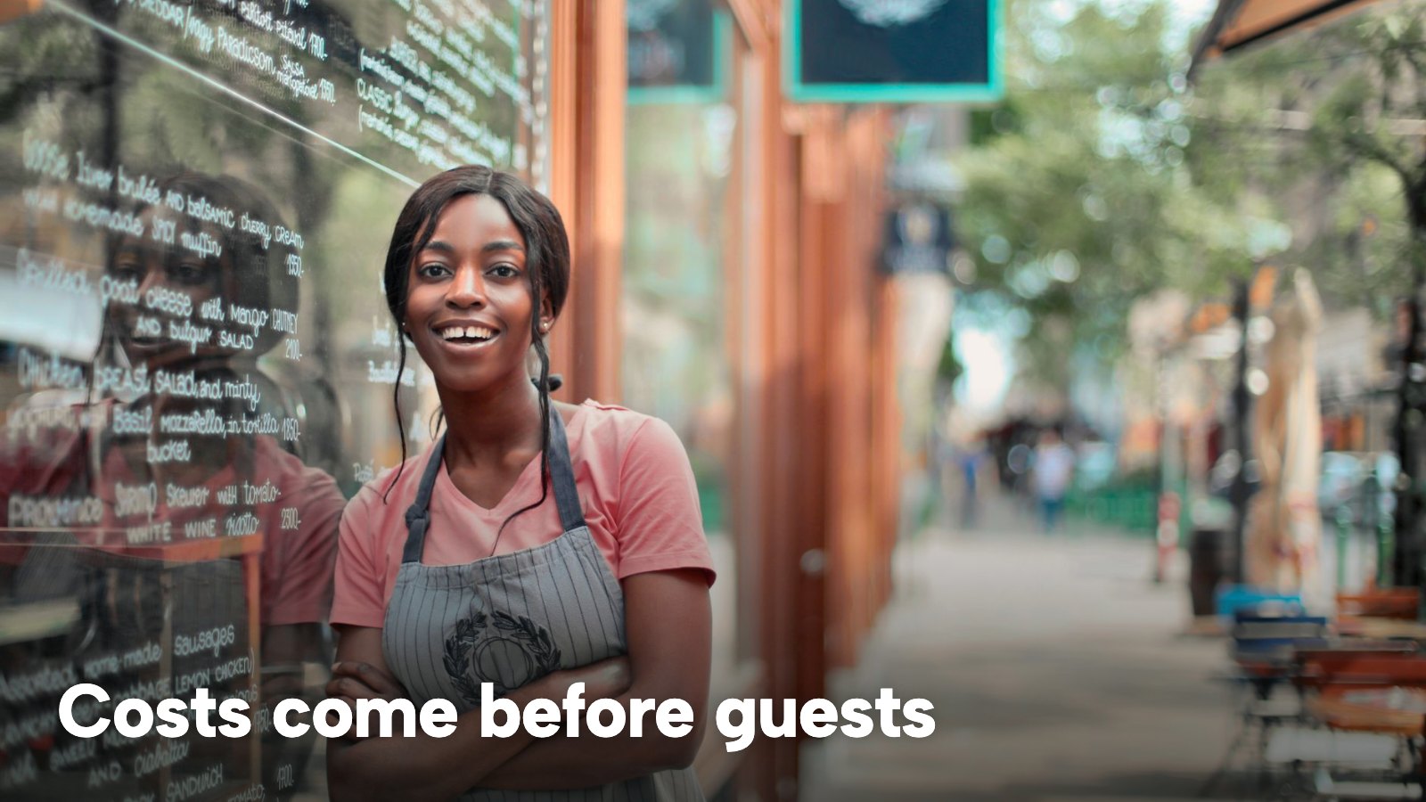 A smiling server in an apron stands outside a restaurant on a sunny street. The image carries the words "Costs come before guests".