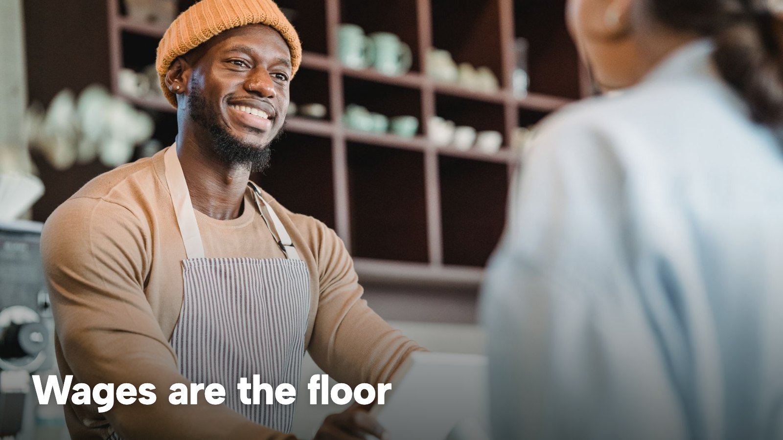 A smiling barista takes an order from a customer at a café counter. The image carries the words "Wages are the floor".