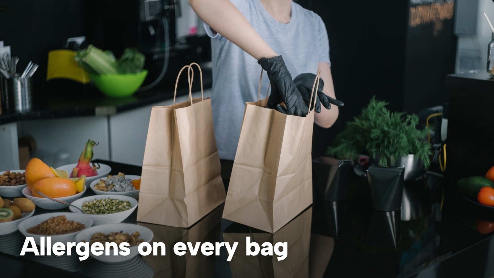 A kitchen worker packs takeaway containers into paper bags. The image carries the words "Allergens on every bag".