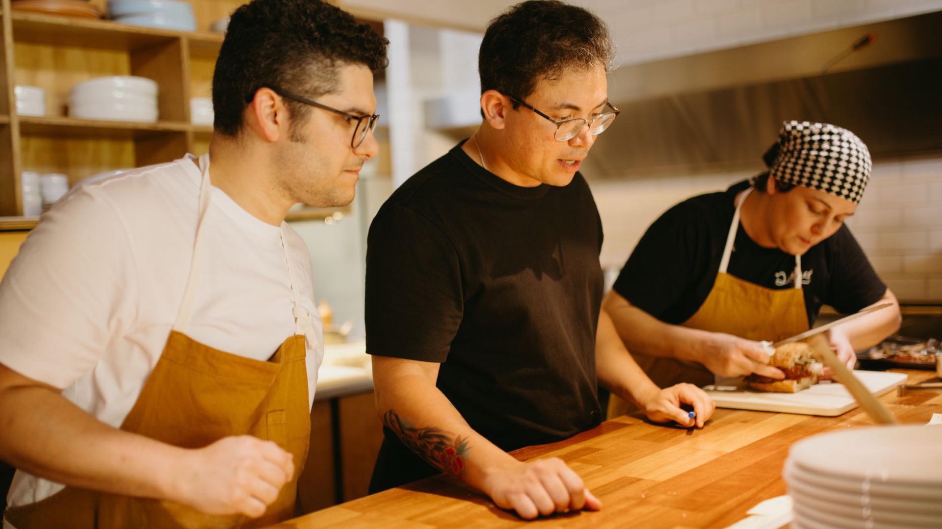 Three cooks preparing food at a wooden pass in a restaurant kitchen.