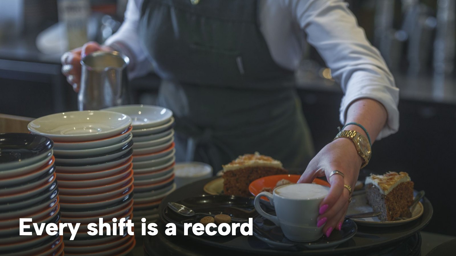 A café worker sets down a coffee and a slice of cake for a guest during a busy service. The image carries the words "Every shift is a record".