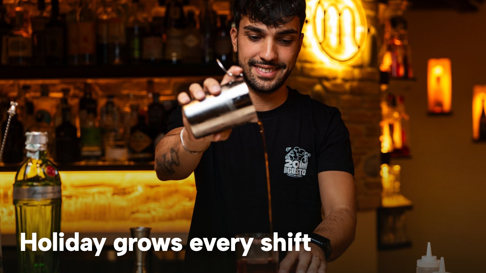 A bartender shakes a cocktail behind a lit bar. The image carries the words "Holiday grows every shift".