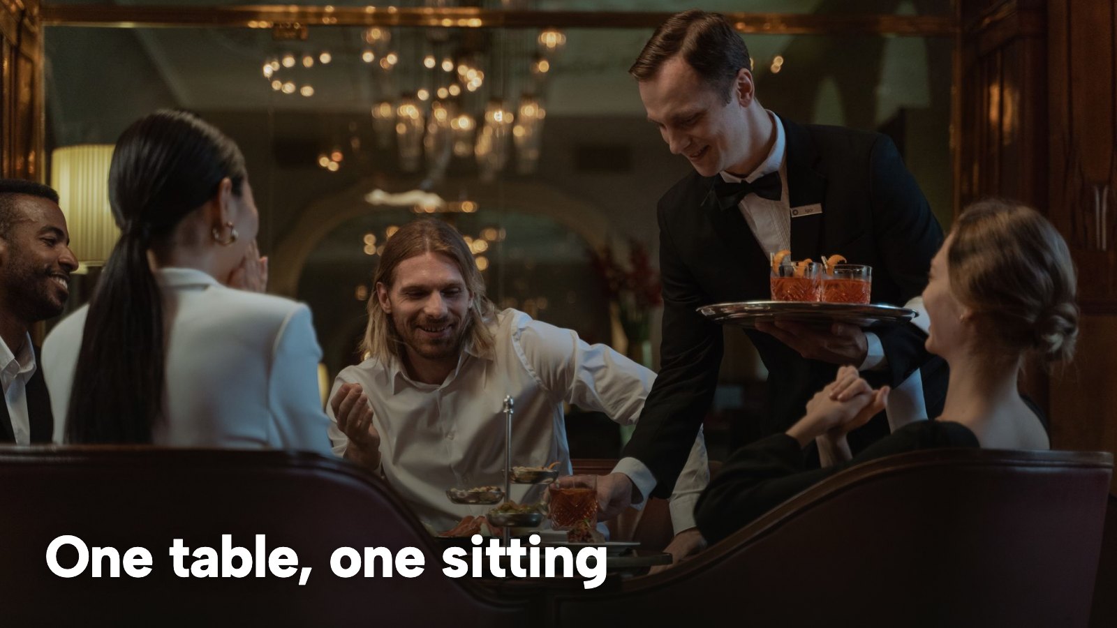 A waiter serving appetisers to a group of diners in a formal dining room. The image carries the words "One table, one sitting".