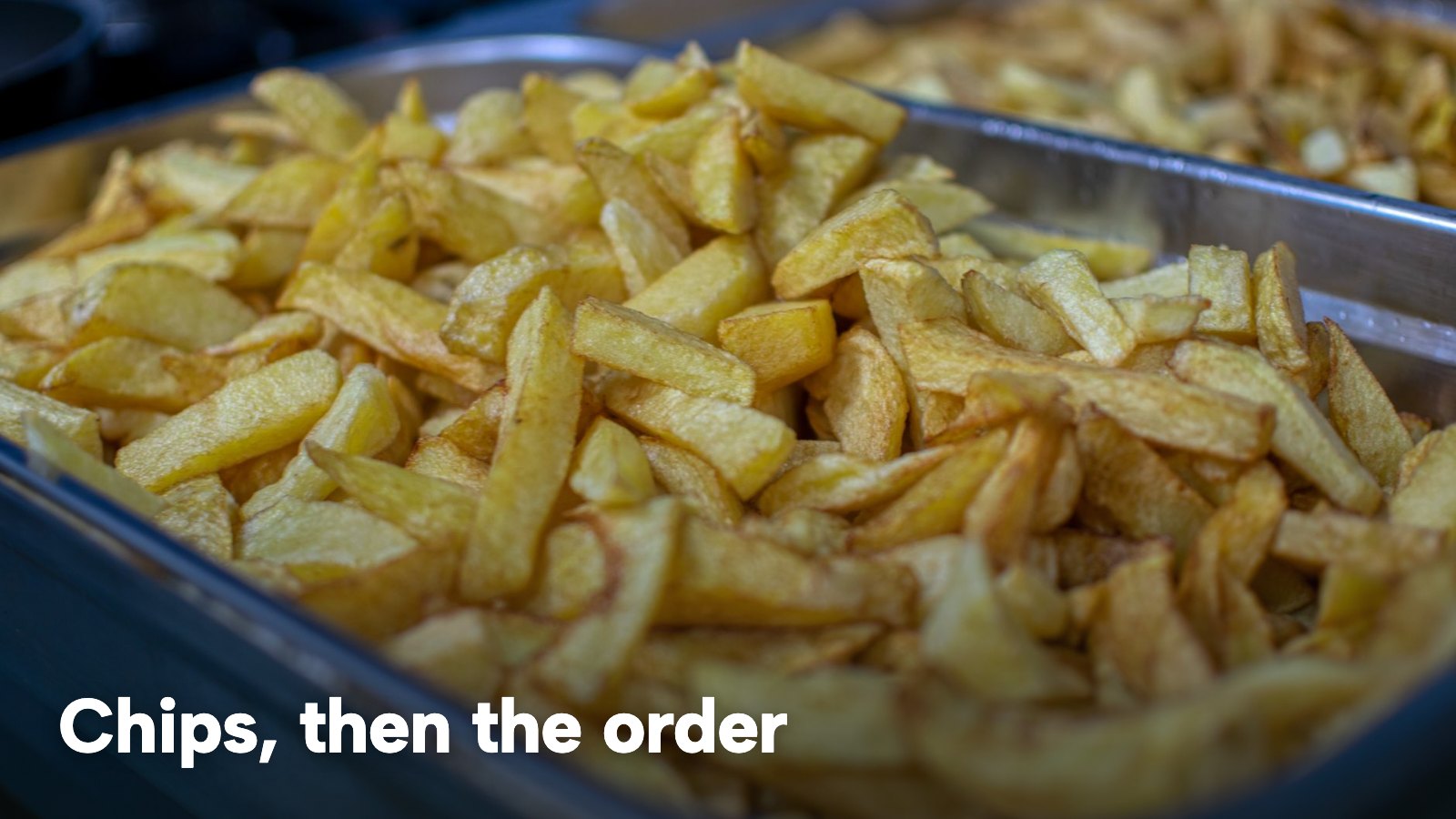 A metal tray of golden chips under the lights of a takeaway counter. The image carries the words "Chips, then the order".