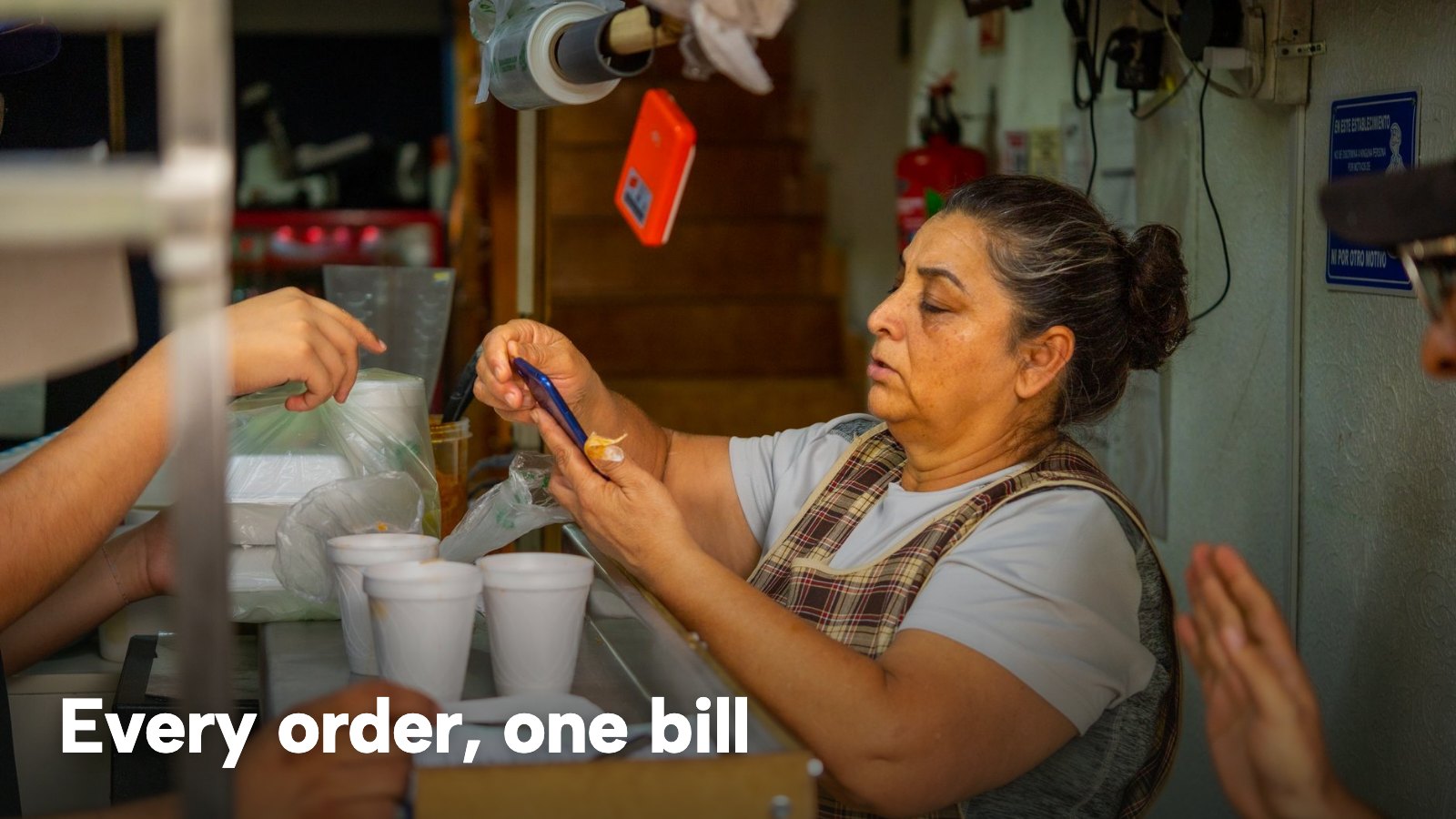 A woman serving customers at a busy food counter while holding a smartphone. The image carries the words "Every order, one bill".