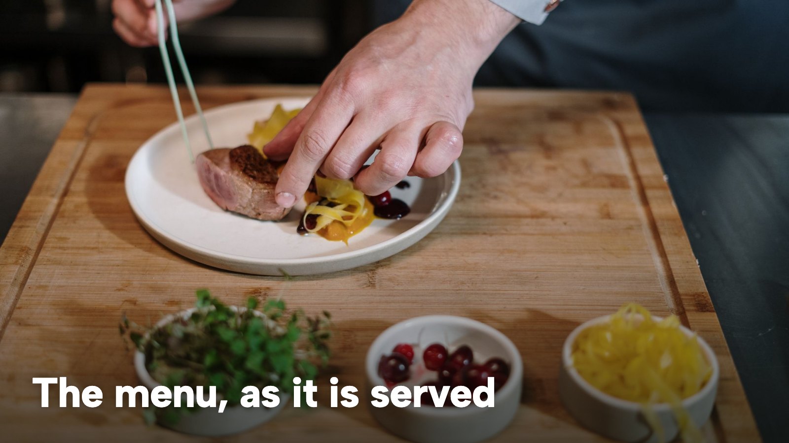 A chef carefully plating a dish in a restaurant kitchen. The image carries the words "The menu, as it is served".