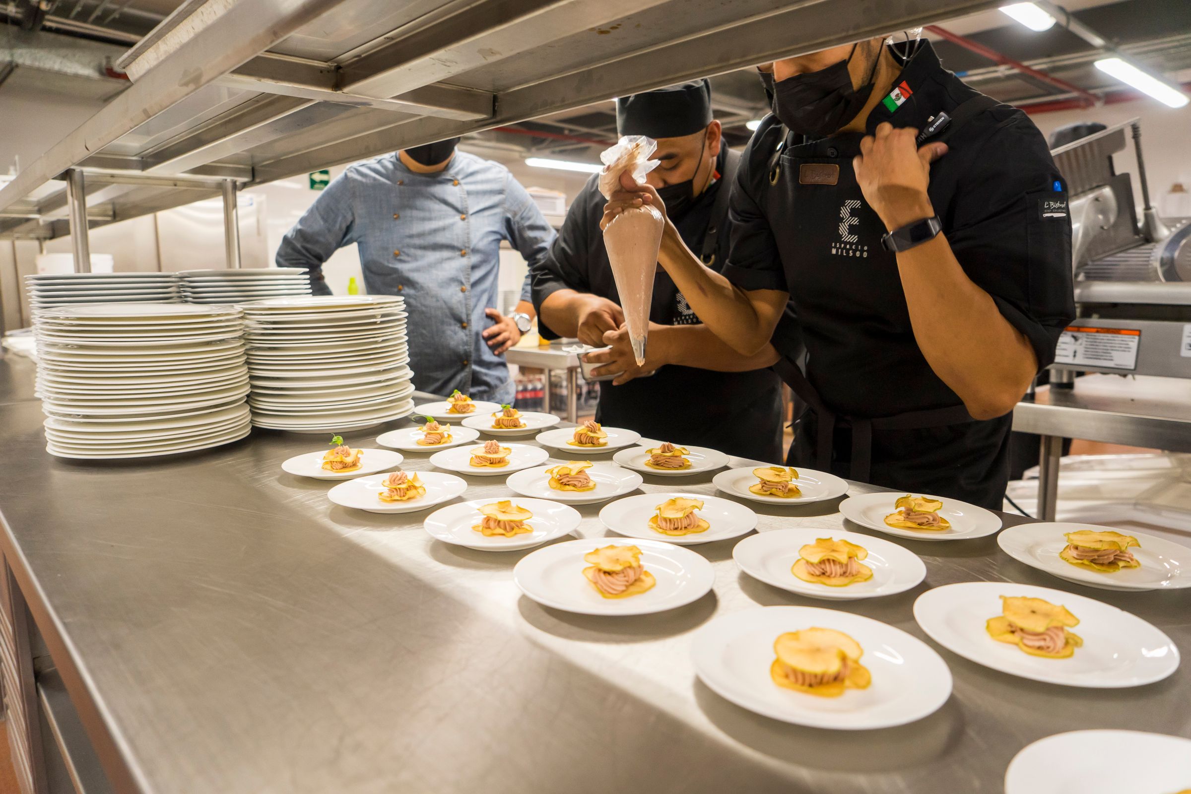 A row of plated dishes lined up on a stainless-steel pass while chefs finish them.