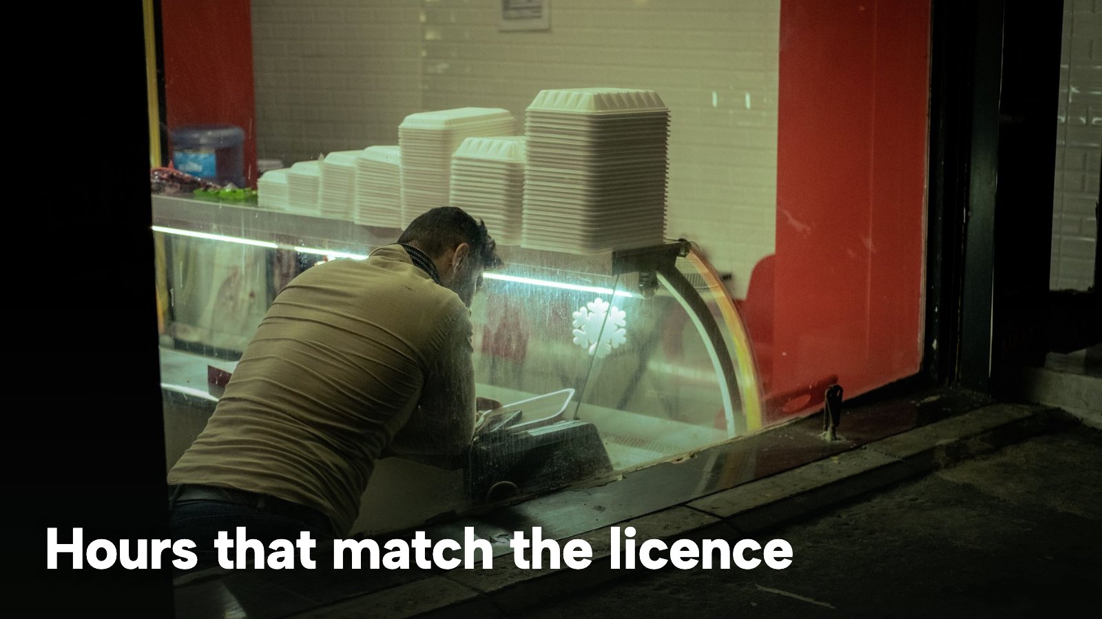 A man arranging food containers in a takeaway at night, seen through the window. The image carries the words "Hours that match the licence".