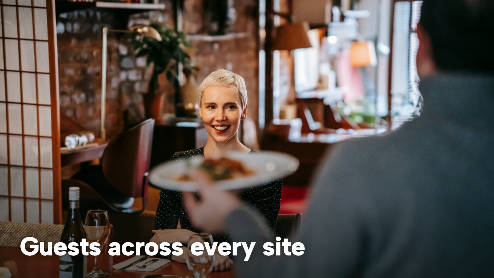 A server carries a plate to a smiling guest seated in a busy restaurant. The image carries the words "Guests across every site".