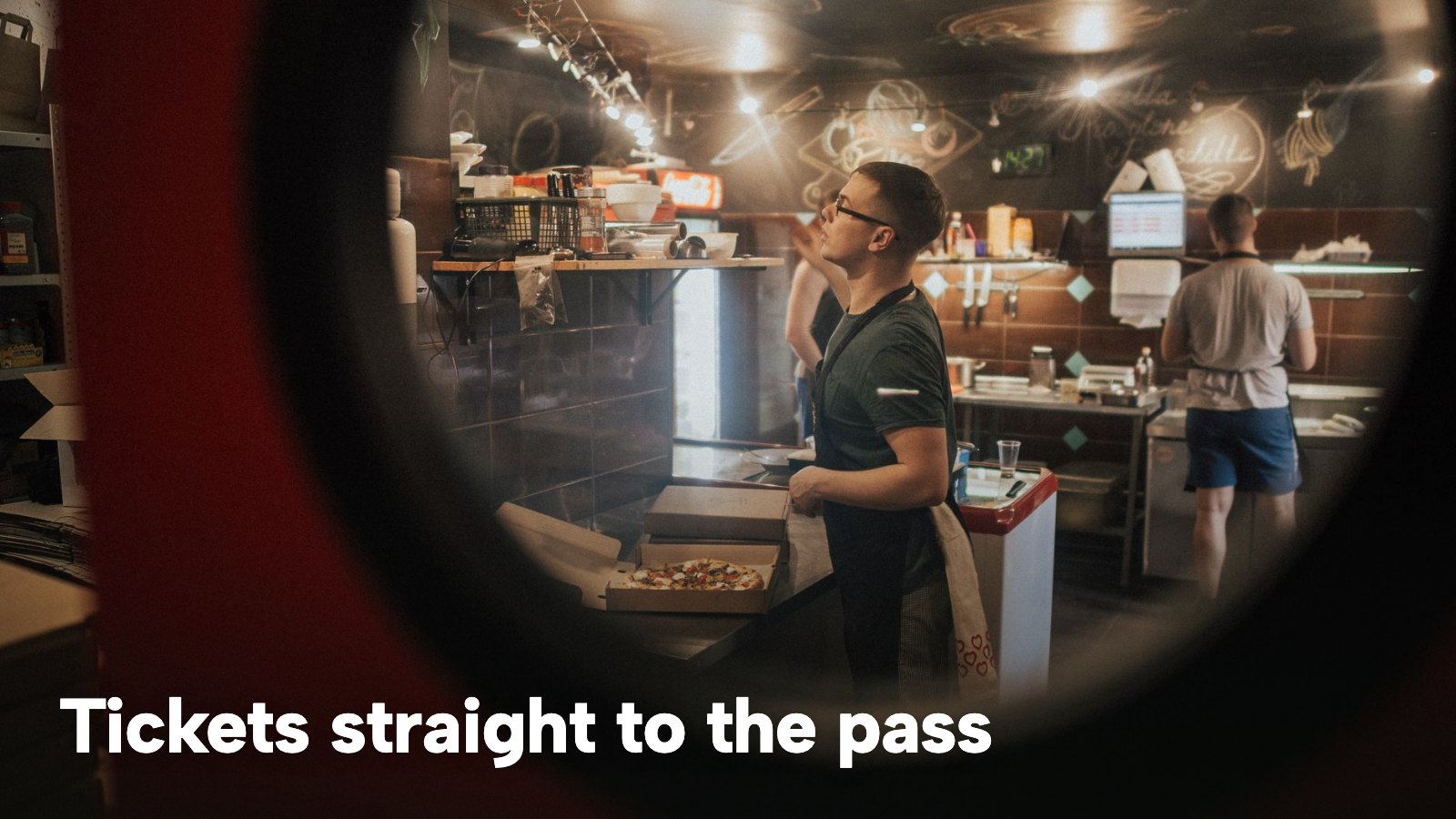 A pizzeria kitchen seen through a round window, with staff at work and pizza boxes stacked. The image carries the words "Tickets straight to the pass".