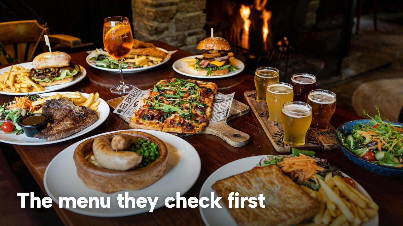 A table of pub food and drinks beside a fireplace. The image carries the words "The menu they check first".