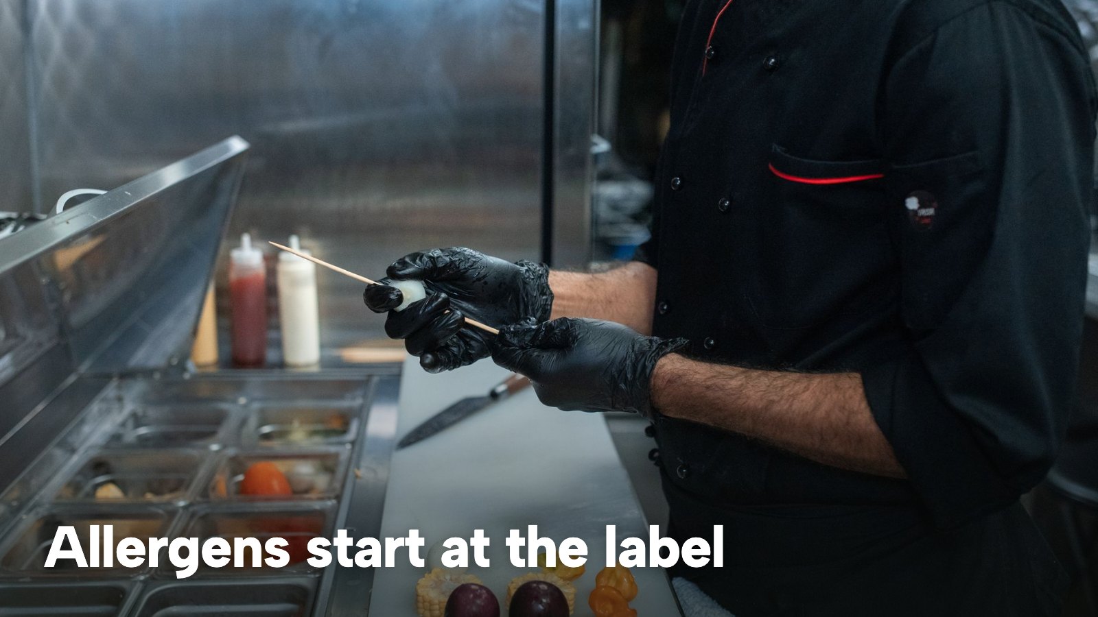 A cook in black gloves taking ingredients from labelled prep containers in a restaurant kitchen. The image carries the words "Allergens start at the label".