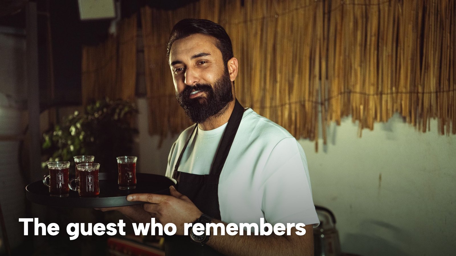 A smiling server carries a tray of drinks across a small restaurant. The image carries the words "The guest who remembers".