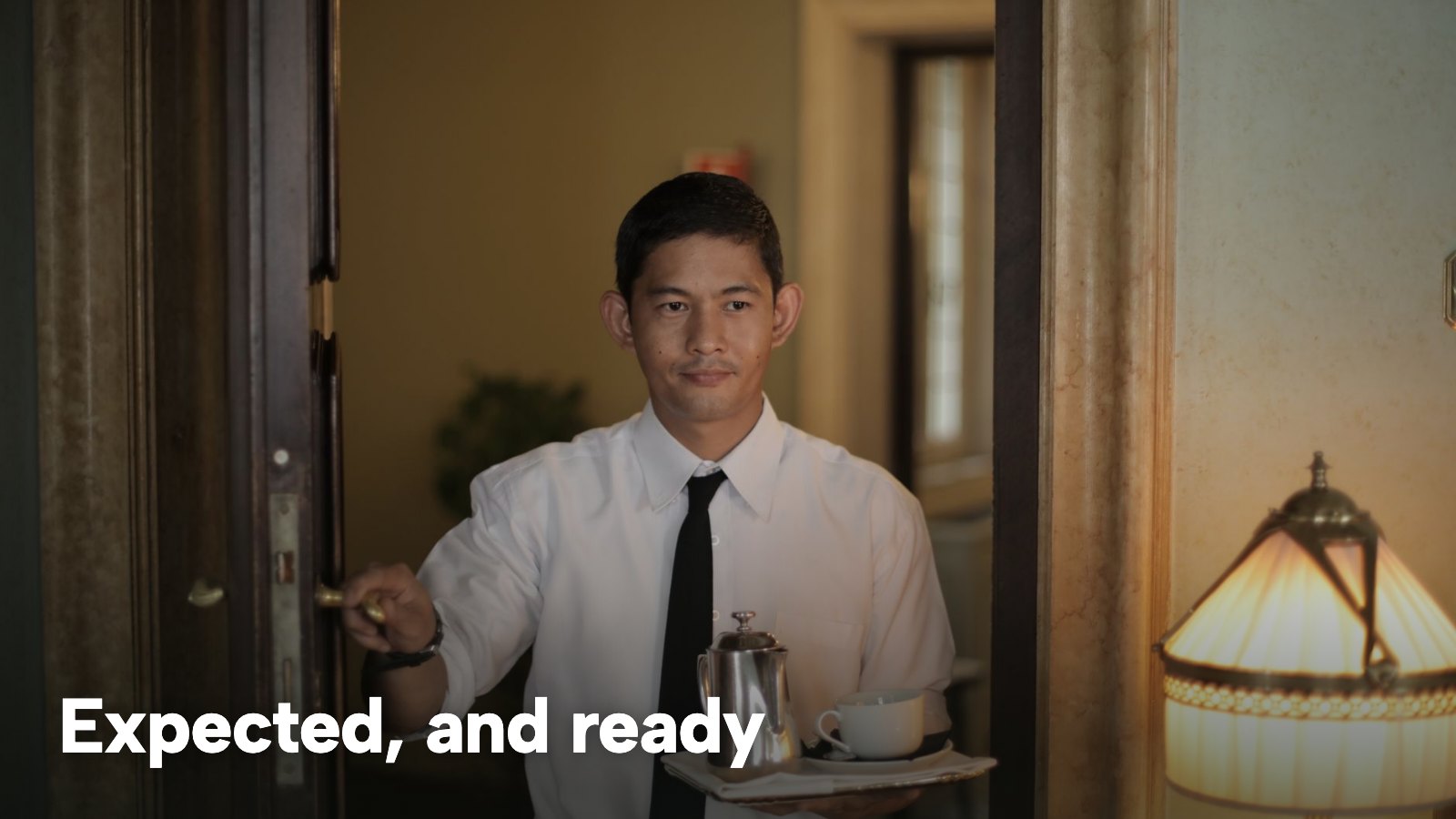 A server in a white shirt and tie opens a door, holding a tray with a coffee pot. The image carries the words "Expected, and ready".