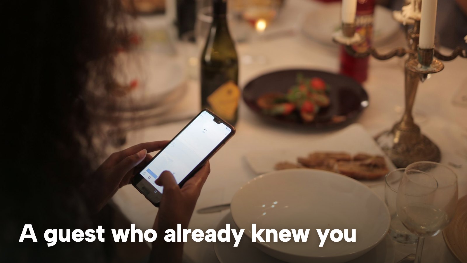 A diner holds a phone at a candlelit restaurant table set with plates and glasses. The image carries the words "A guest who already knew you".
