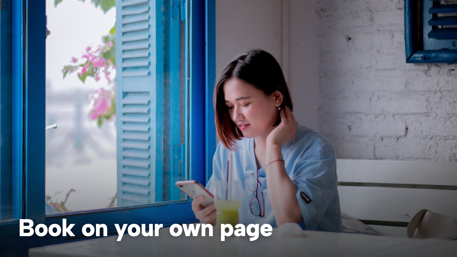 A woman sits at a café table by a blue-framed window, looking at her phone. The image carries the words "Book on your own page".