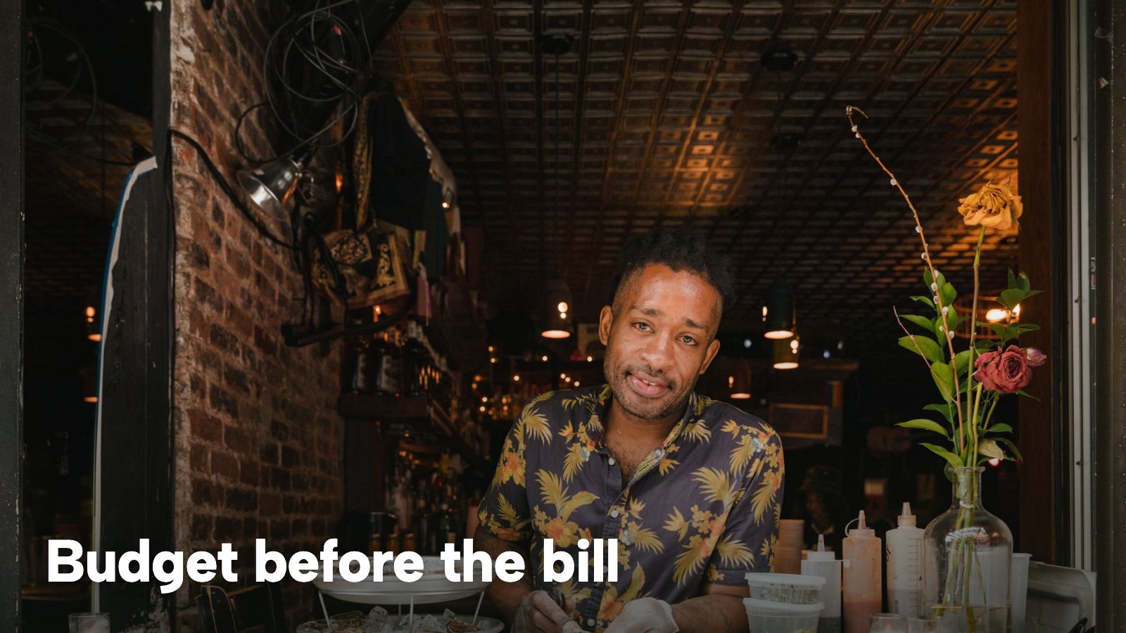 A smiling owner in a patterned shirt preparing food at a small restaurant counter. The image carries the words "Budget before the bill".