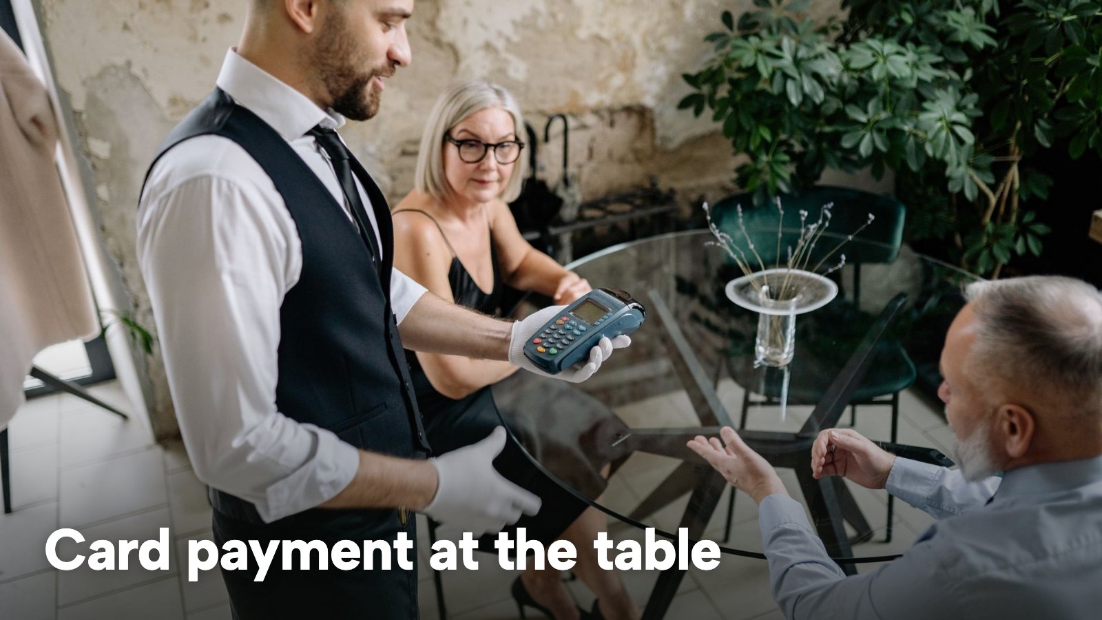A waiter holding a card terminal beside a seated couple at a glass table in a restaurant. The image carries the words "Card payment at the table".