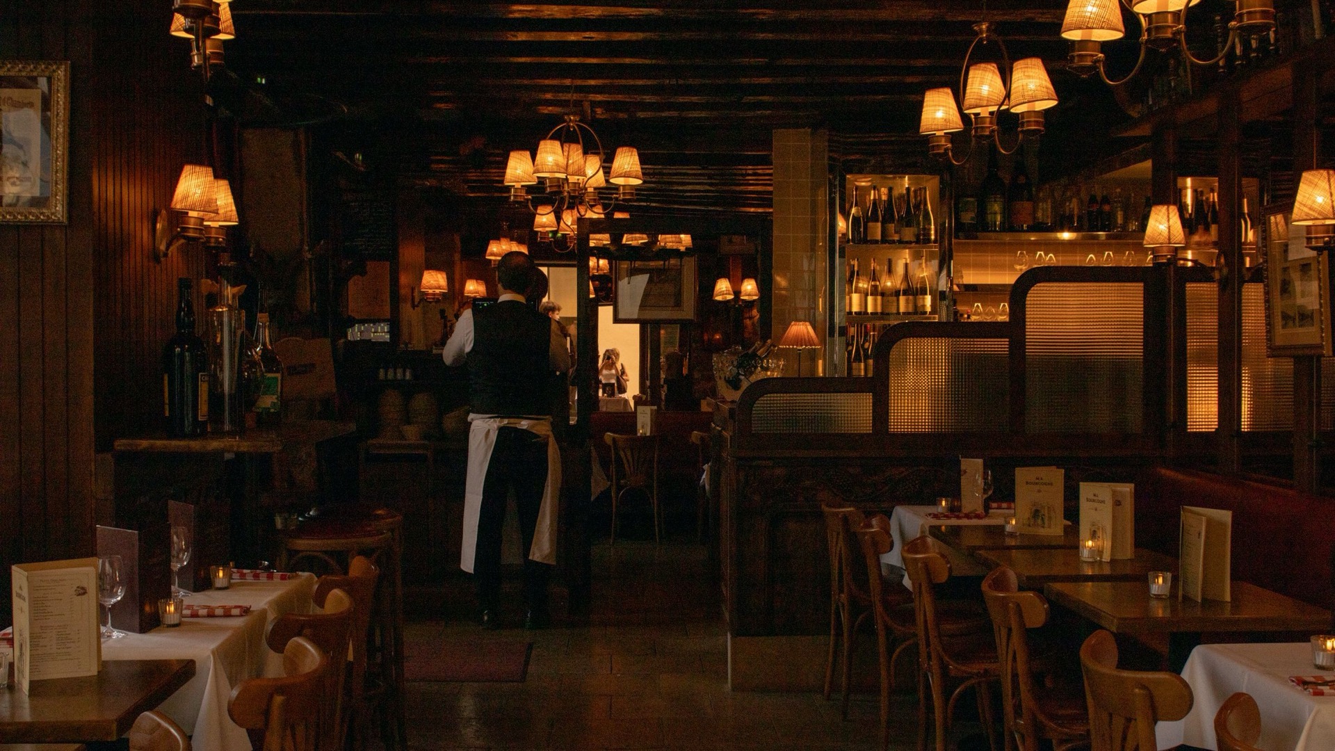 A dimly lit restaurant dining room with wooden decor and a waiter at work.