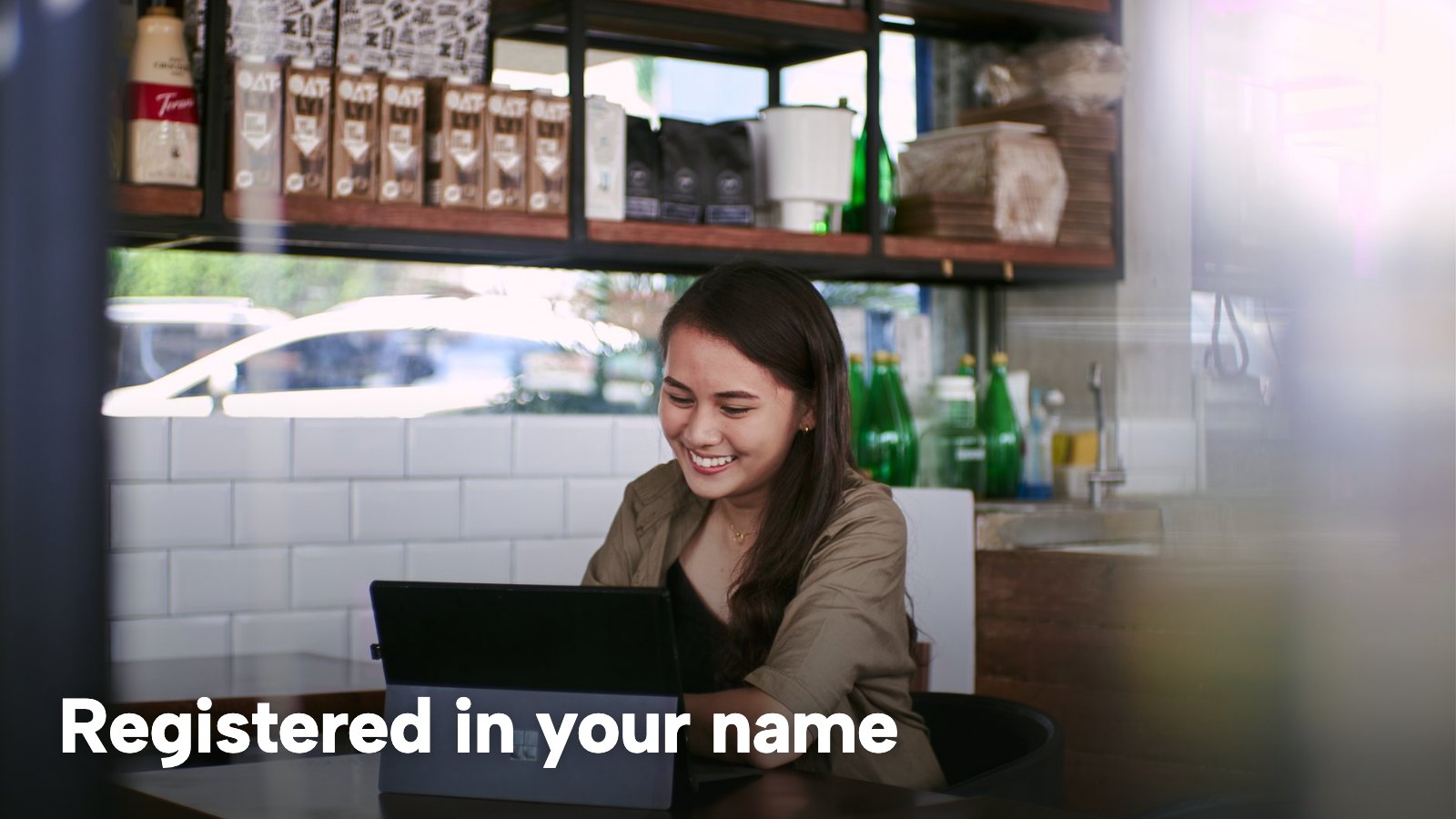 A smiling woman works on a laptop in a café. The image carries the words "Registered in your name".