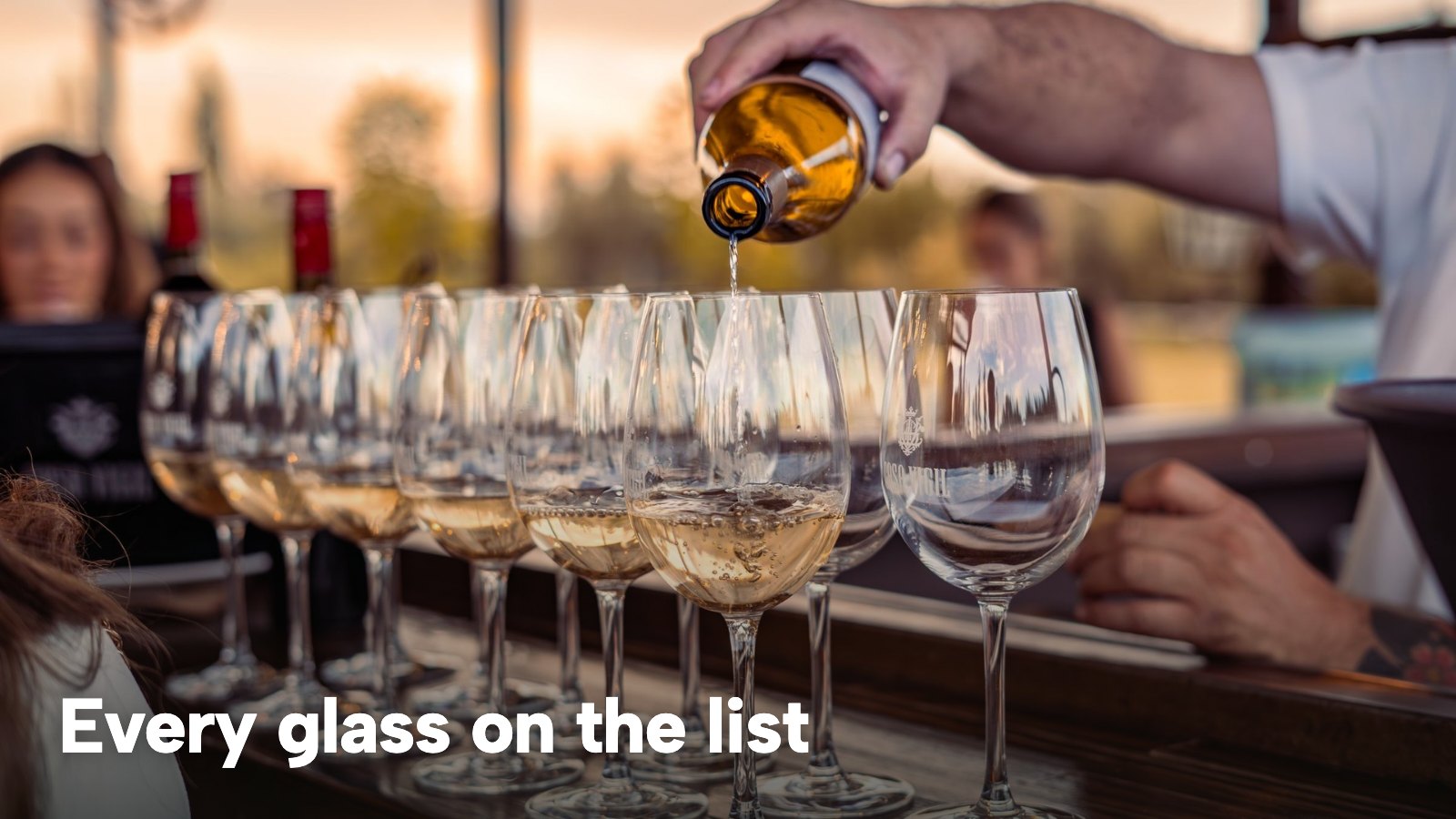 A bartender pouring white wine into a row of glasses on a bar. The image carries the words "Every glass on the list".