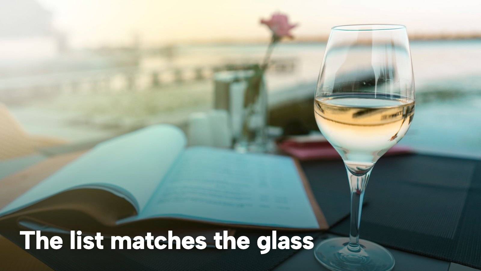 A glass of white wine beside an open menu on a table by the water. The image carries the words "The list matches the glass".
