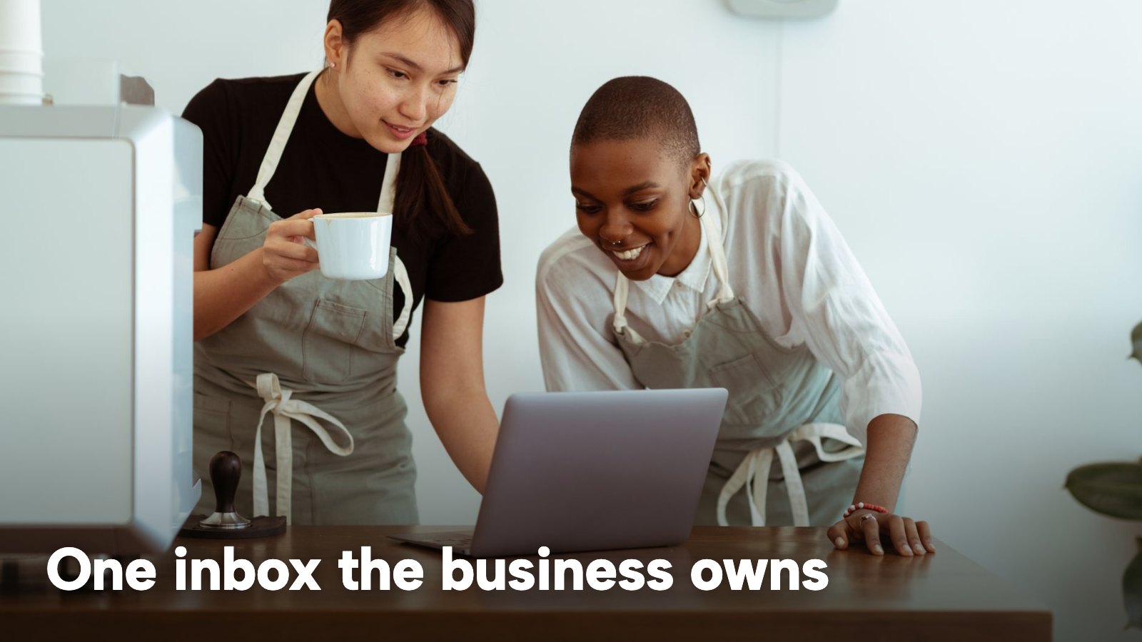 Two members of a café team looking at a laptop together at the counter. The image carries the words "One inbox the business owns".