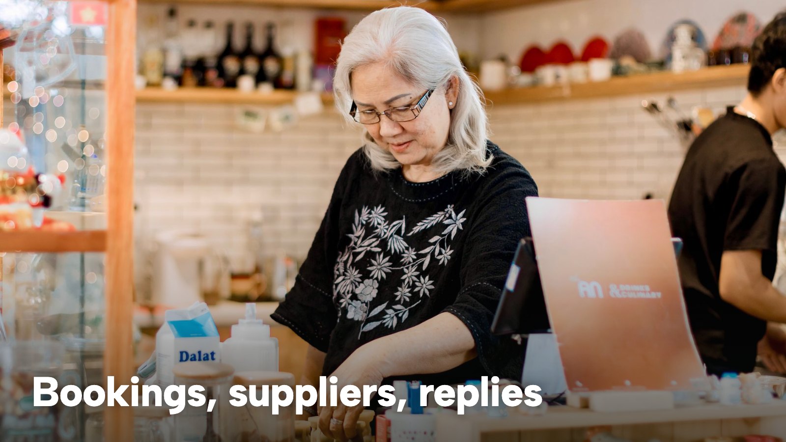 A café owner at the counter working on a laptop beside the till. The image carries the words "Bookings, suppliers, replies".