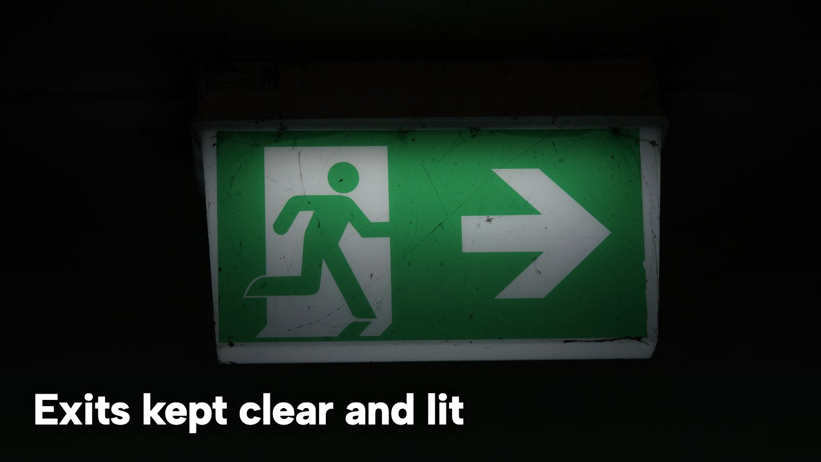 An illuminated green emergency exit sign with an arrow, in a dark corridor. The image carries the words "Exits kept clear and lit".