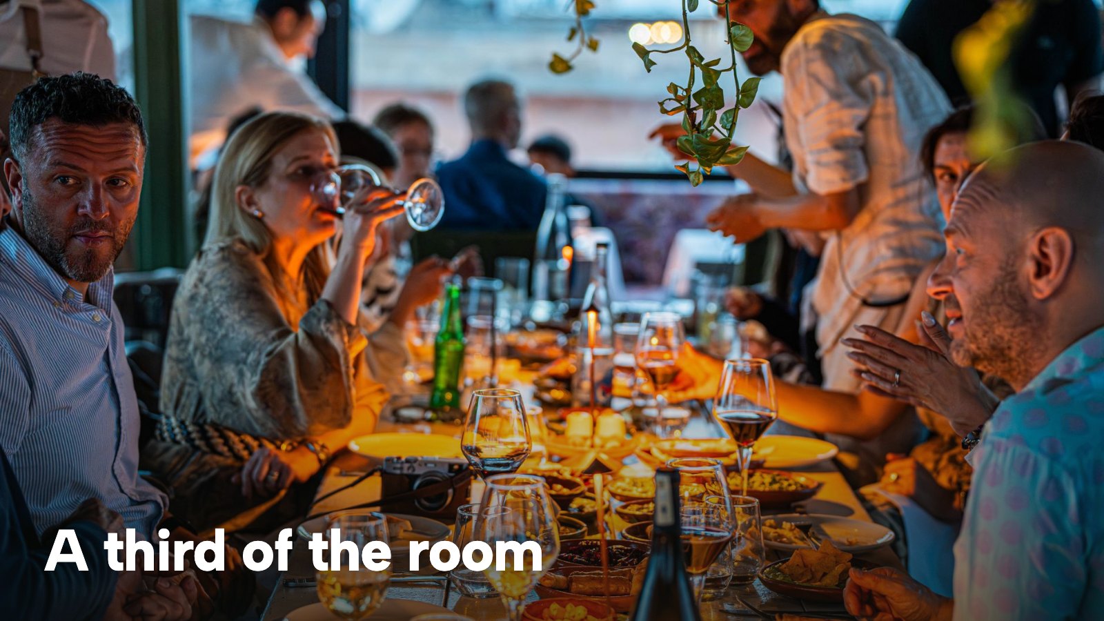 A large group of friends share a candlelit dinner along a long restaurant table. The image carries the words "A third of the room".