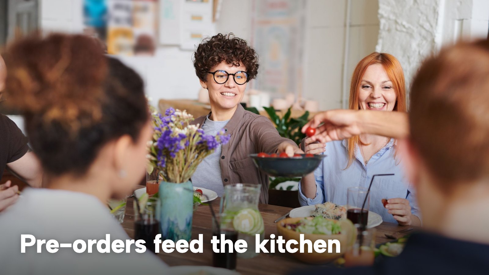 A group of friends and family enjoy a shared meal at a table indoors. The image carries the words "Pre-orders feed the kitchen".