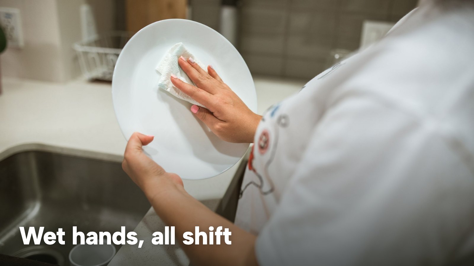A person cleaning a ceramic plate by hand at a kitchen sink. The image carries the words "Wet hands, all shift".