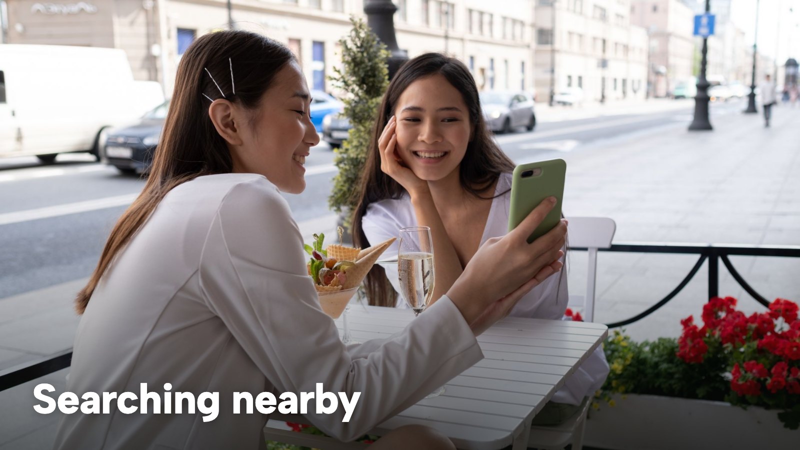 Two women at an outdoor table look at a phone together. The image carries the words "Searching nearby".