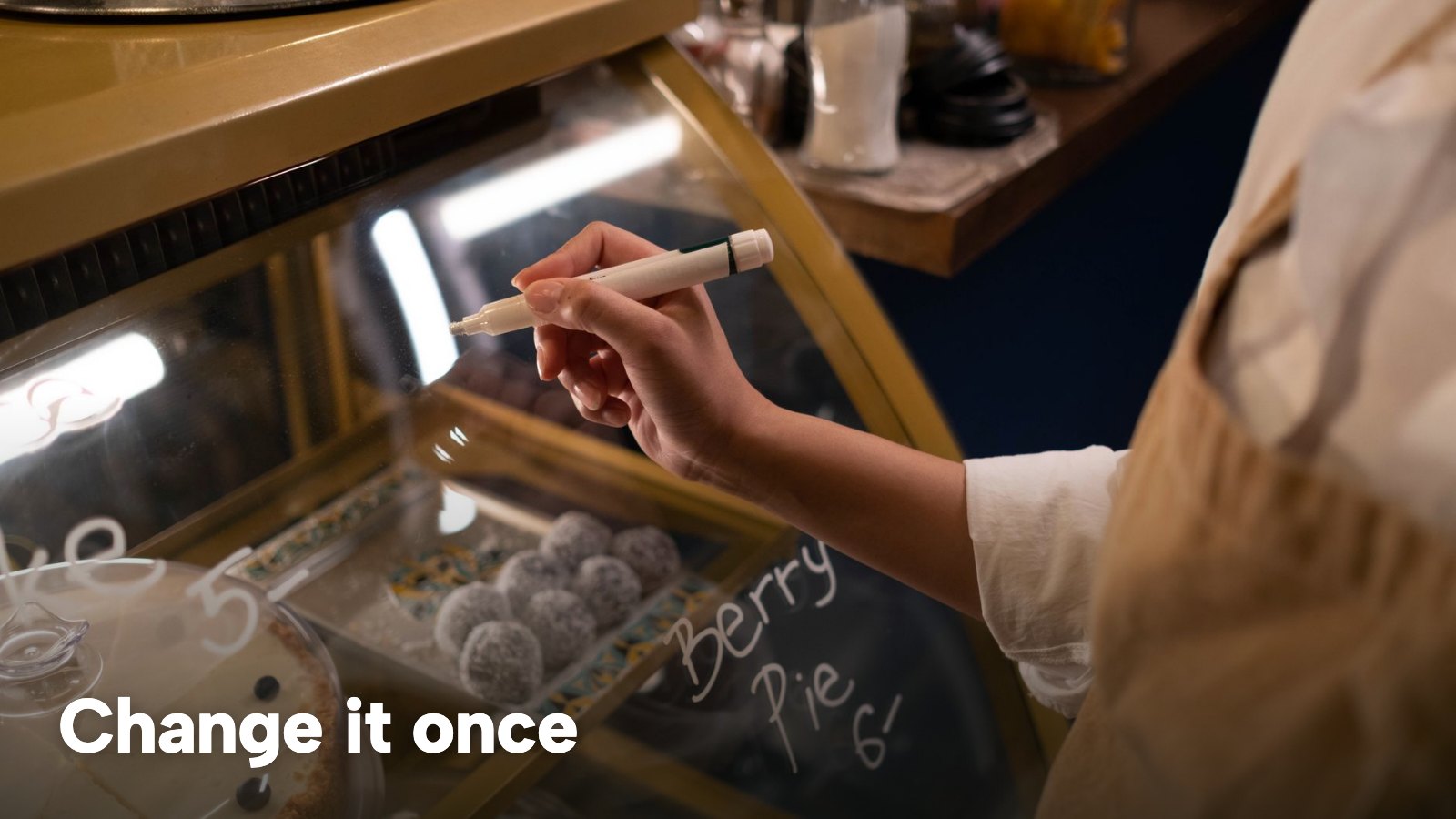 A hand writes a dish name and price on a café display sign. The image carries the words "Change it once".