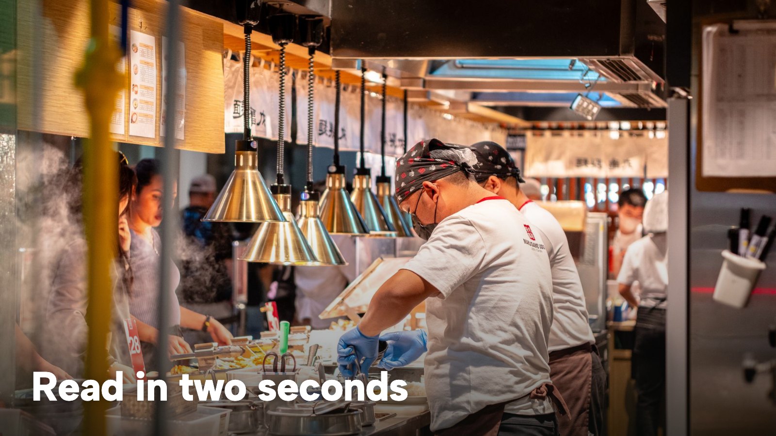 Cooks working under heat lamps along a busy open kitchen line. The image carries the words "Read in two seconds".