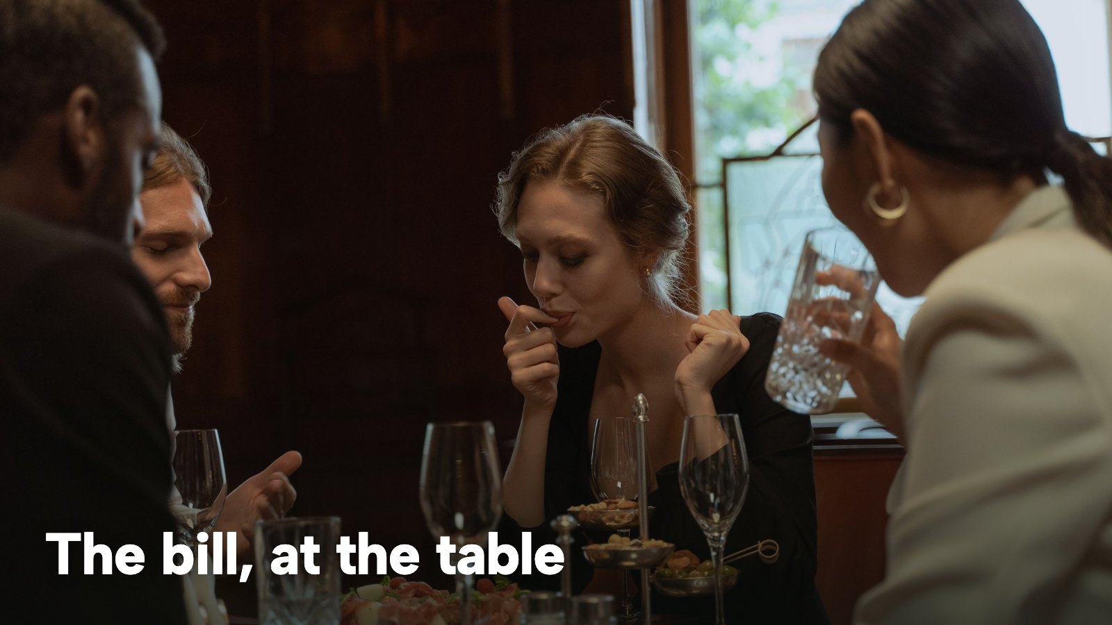Friends at a restaurant dinner with drinks and plates on the table. The image carries the words "The bill, at the table".
