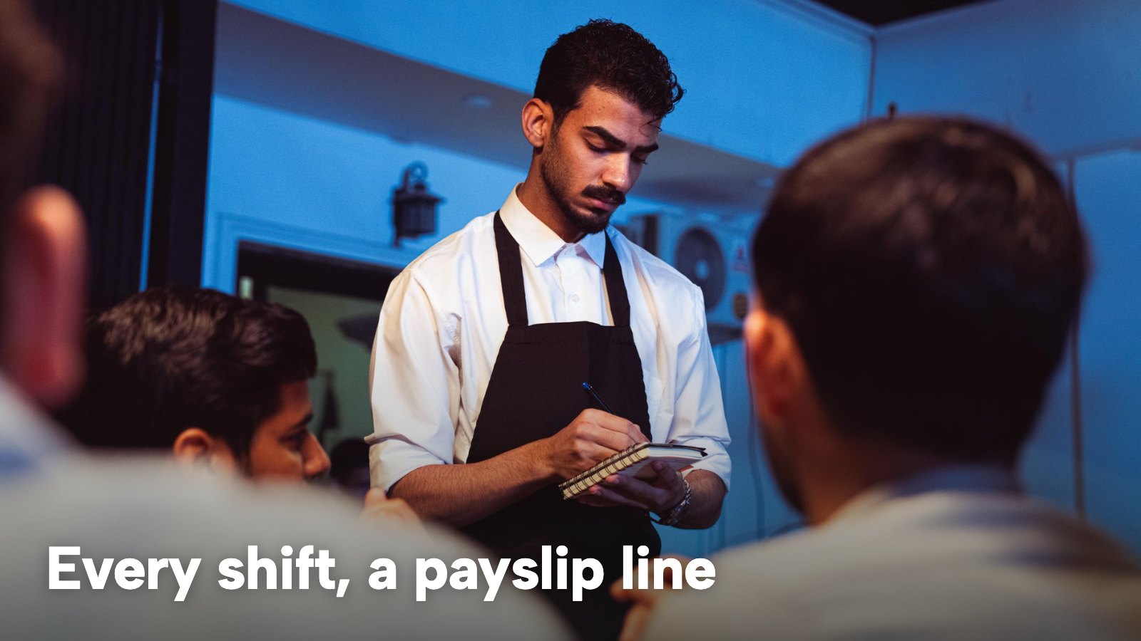 A waiter writes down an order for guests at a restaurant table. The image carries the words "Every shift, a payslip line".