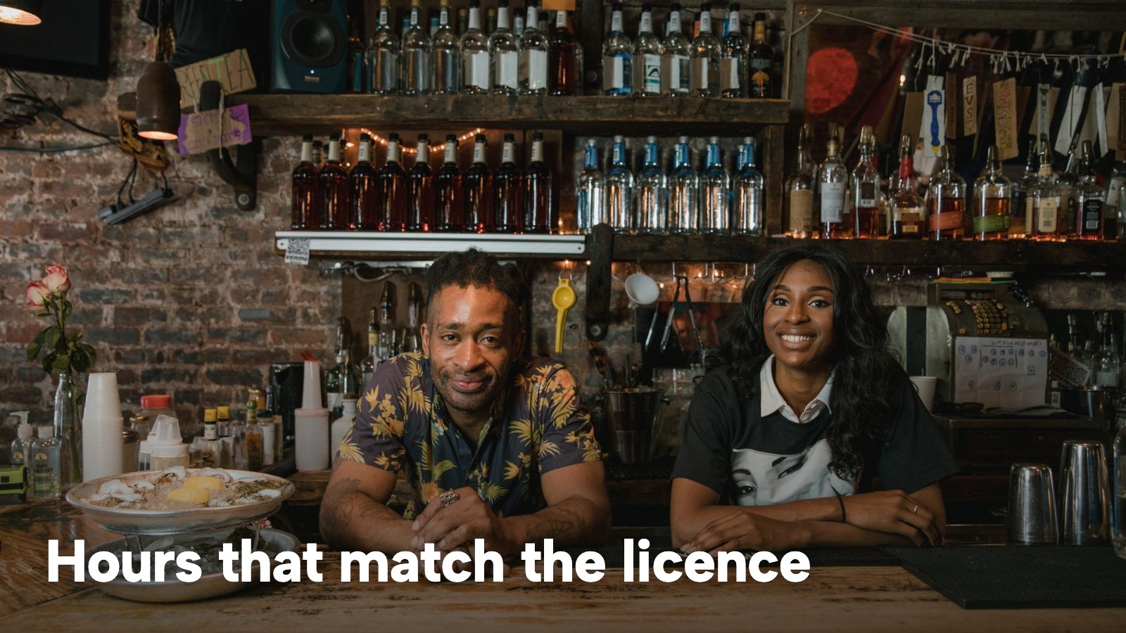 Two smiling bartenders behind a warmly lit bar counter. The image carries the words "Hours that match the licence".