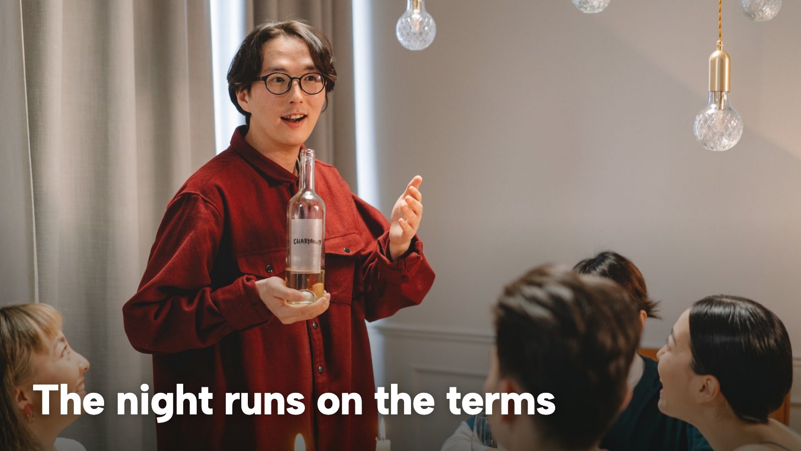 A host holding a bottle of wine and talking to guests seated at a dinner table. The image carries the words "The night runs on the terms".