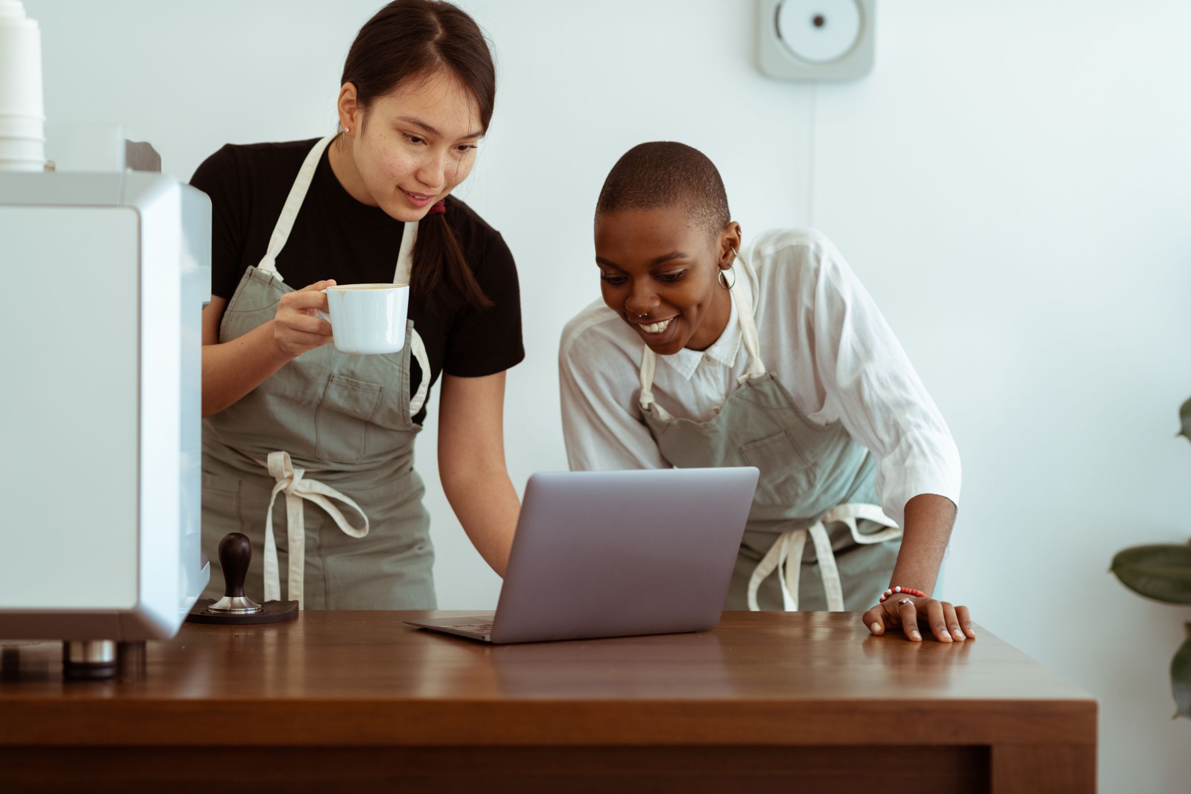 Two café staff in aprons checking a laptop together at the counter.