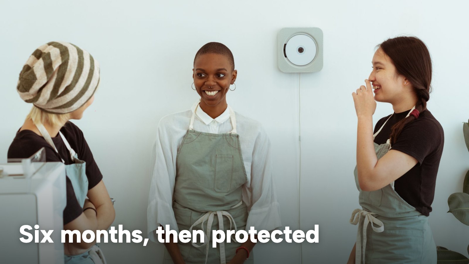 Three young café staff in aprons talk together beside the counter. The image carries the words "Six months, then protected".