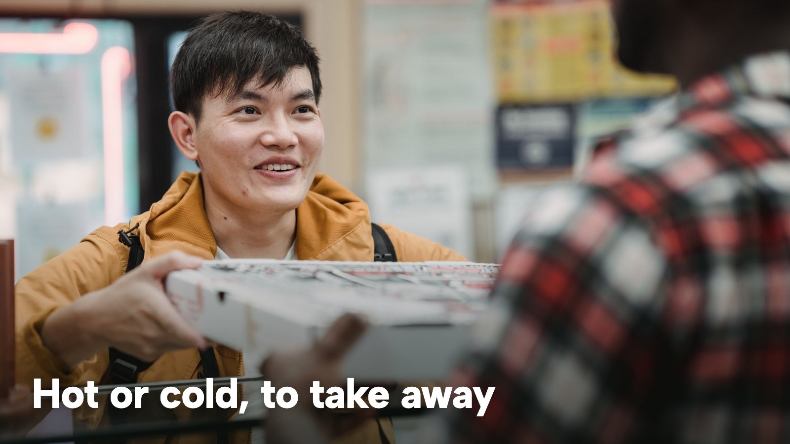 A smiling man collects a takeaway pizza box at a shop counter. The image carries the words "Hot or cold, to take away".