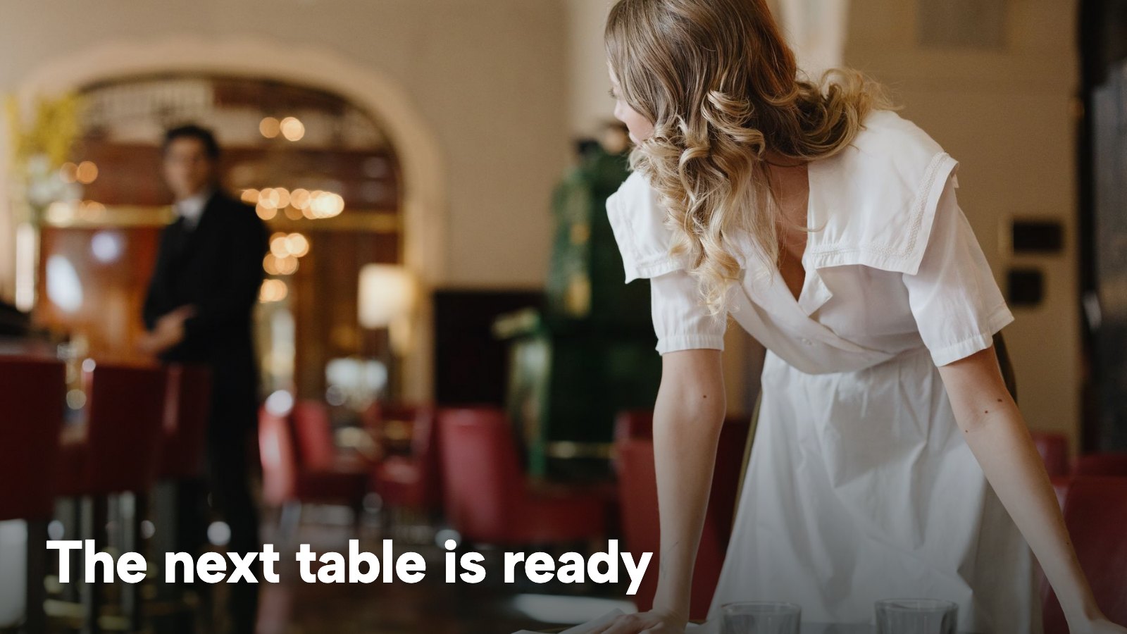 A host in a white dress clearing a table in a dining room as a waiter stands by. The image carries the words "The next table is ready".