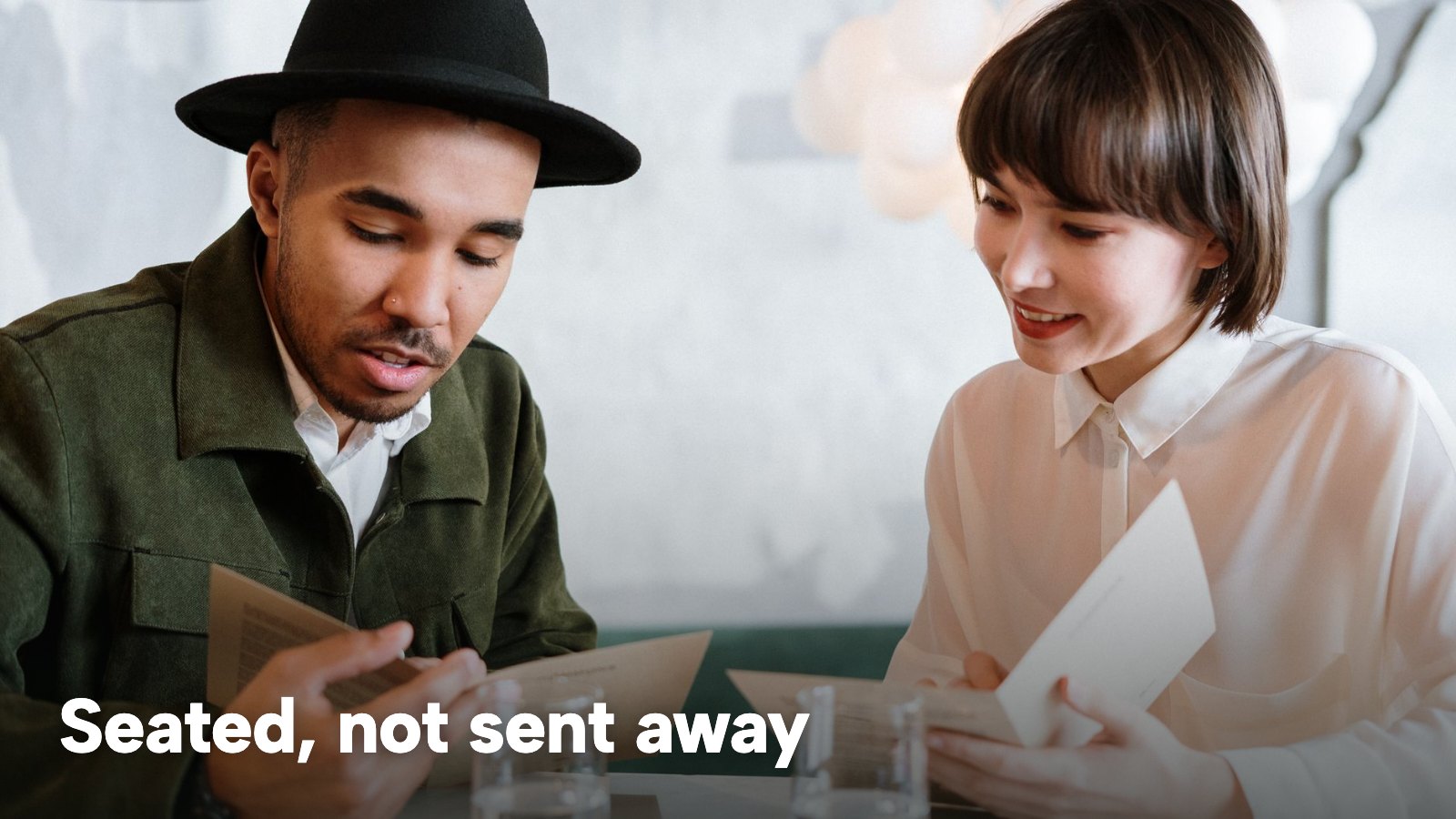 A couple at a café table reading menus together. The image carries the words "Seated, not sent away".