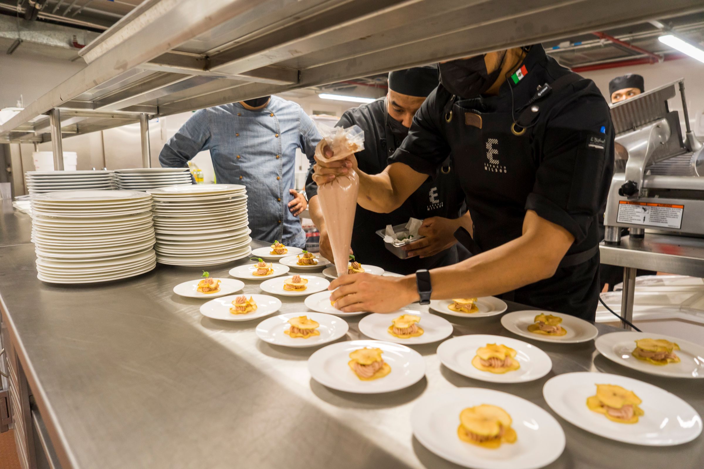 Chefs plating a row of dishes on a stainless-steel pass.